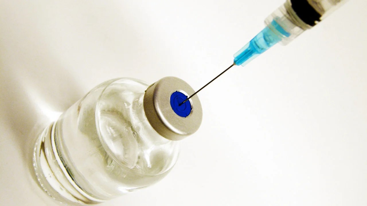 Agulha de uma seringa perfurando a tampa de um frasco-ampola de medicamento injetável, em close