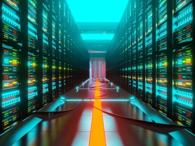 Corredor de um datacenter, com racks de servidores iluminados dos dois lados e cabos no piso central